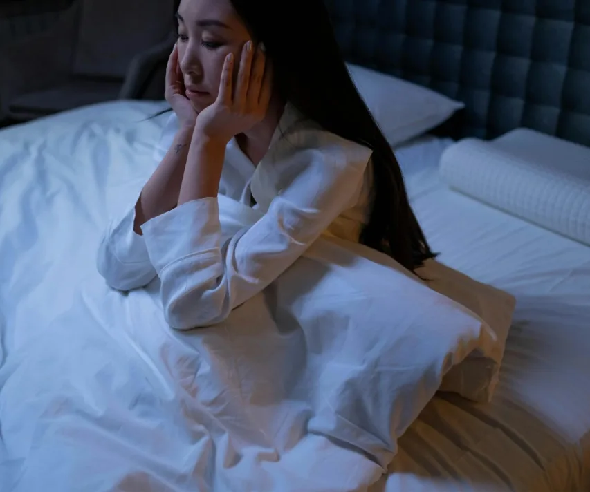 A woman sits on a bed, resting her face in her hands, looking contemplative. The room is dimly lit, creating a serene and quiet atmosphere. She is wearing white pajamas, and the bed is unmade with white sheets and pillows.