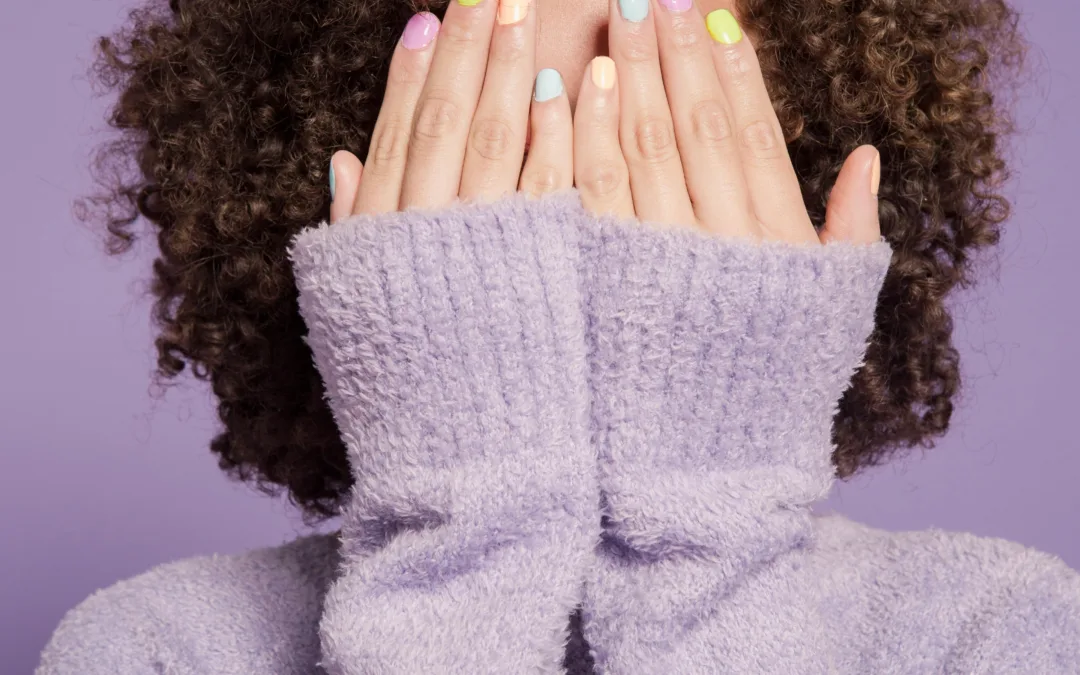 A person with curly hair covers their face with their hands, showing nails painted in pastel colors. They wear a fuzzy, lavender sweater against a purple background.