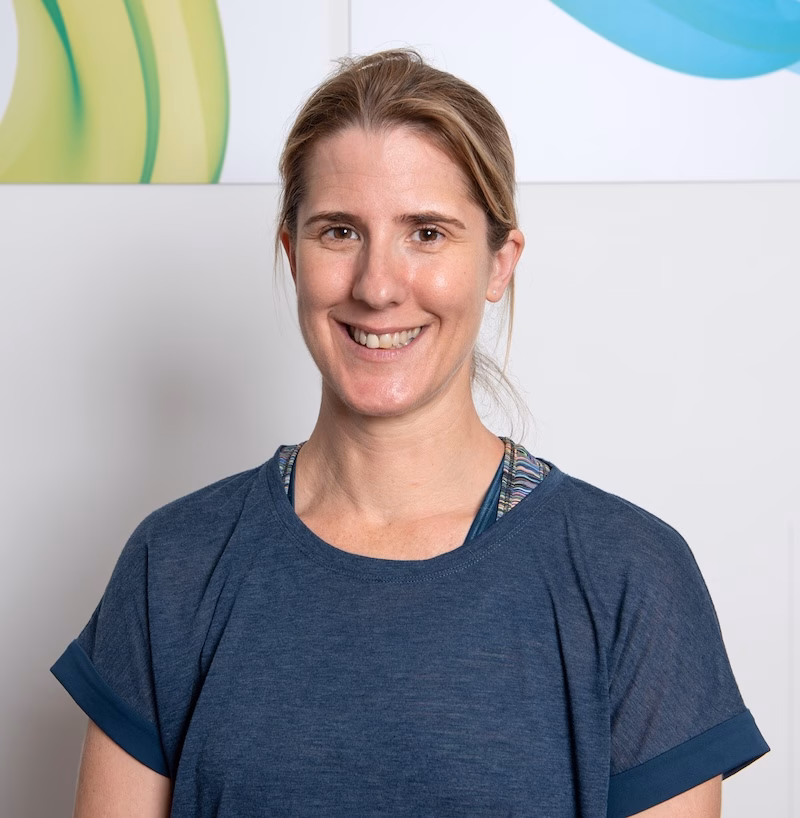 A woman with light brown hair tied back smiles at the camera. She is wearing a blue top and stands indoors against a light-colored wall with abstract art in the background.