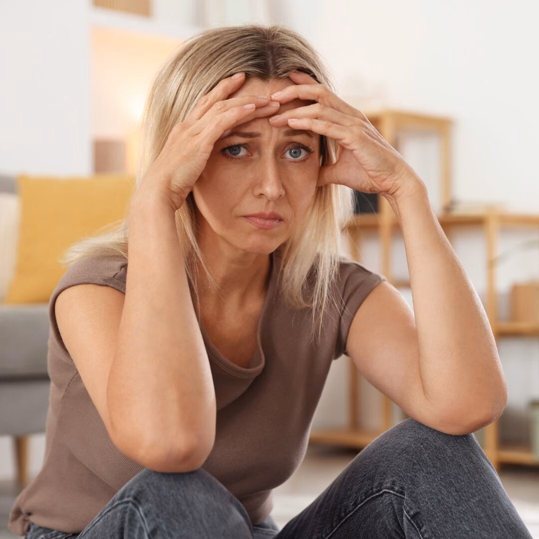 A woman with blonde hair sits indoors, leaning forward with her elbows on her knees and her hands on her forehead, looking concerned or stressed. Shelves and a sofa are visible in the background.